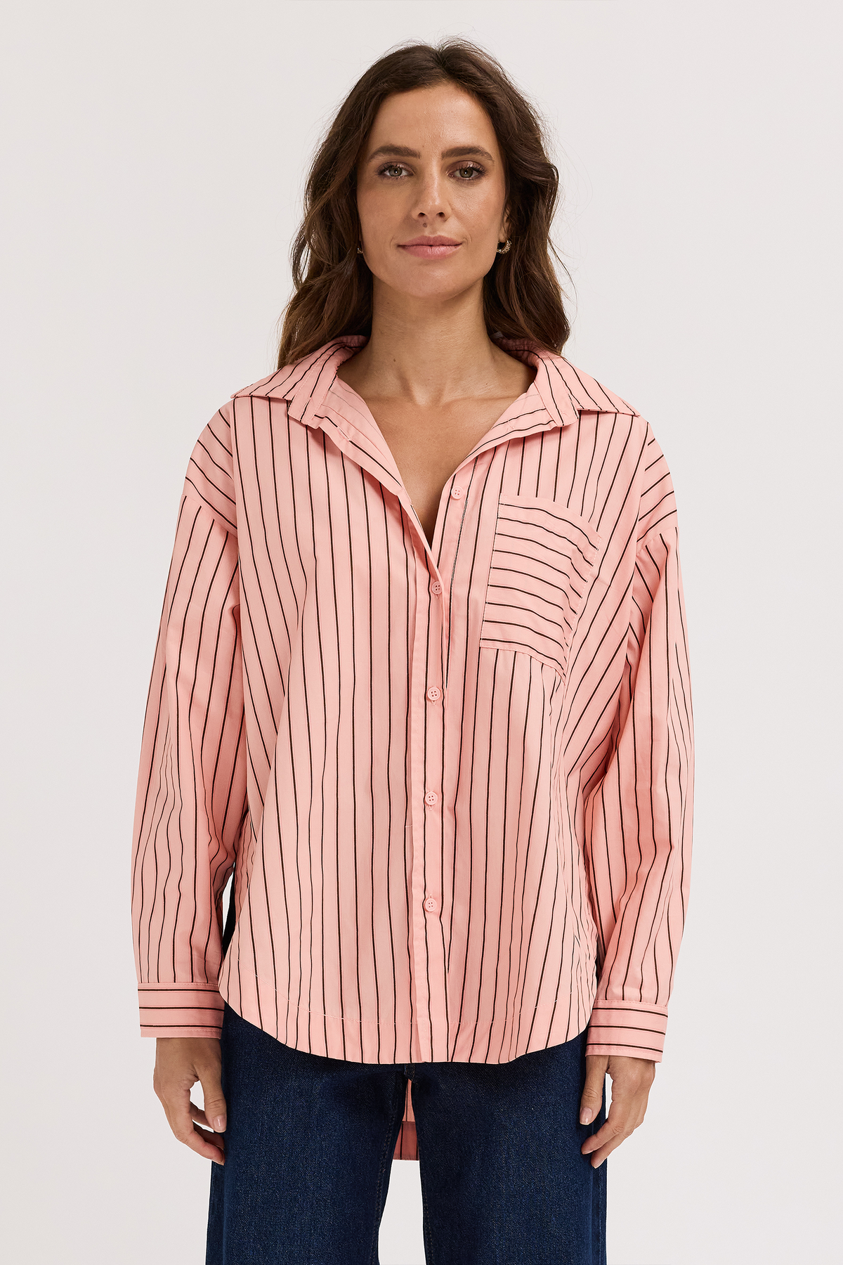 A woman wearing a pink and white vertically striped button-up shirt with a collar and chest pocket, paired with dark blue jeans, against a plain light gray background.