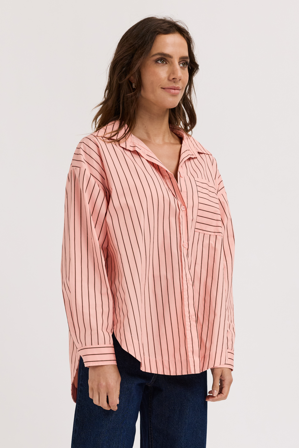 A woman wearing a pink and white vertically striped button-up shirt with dark blue jeans, photographed against a plain light gray studio background.