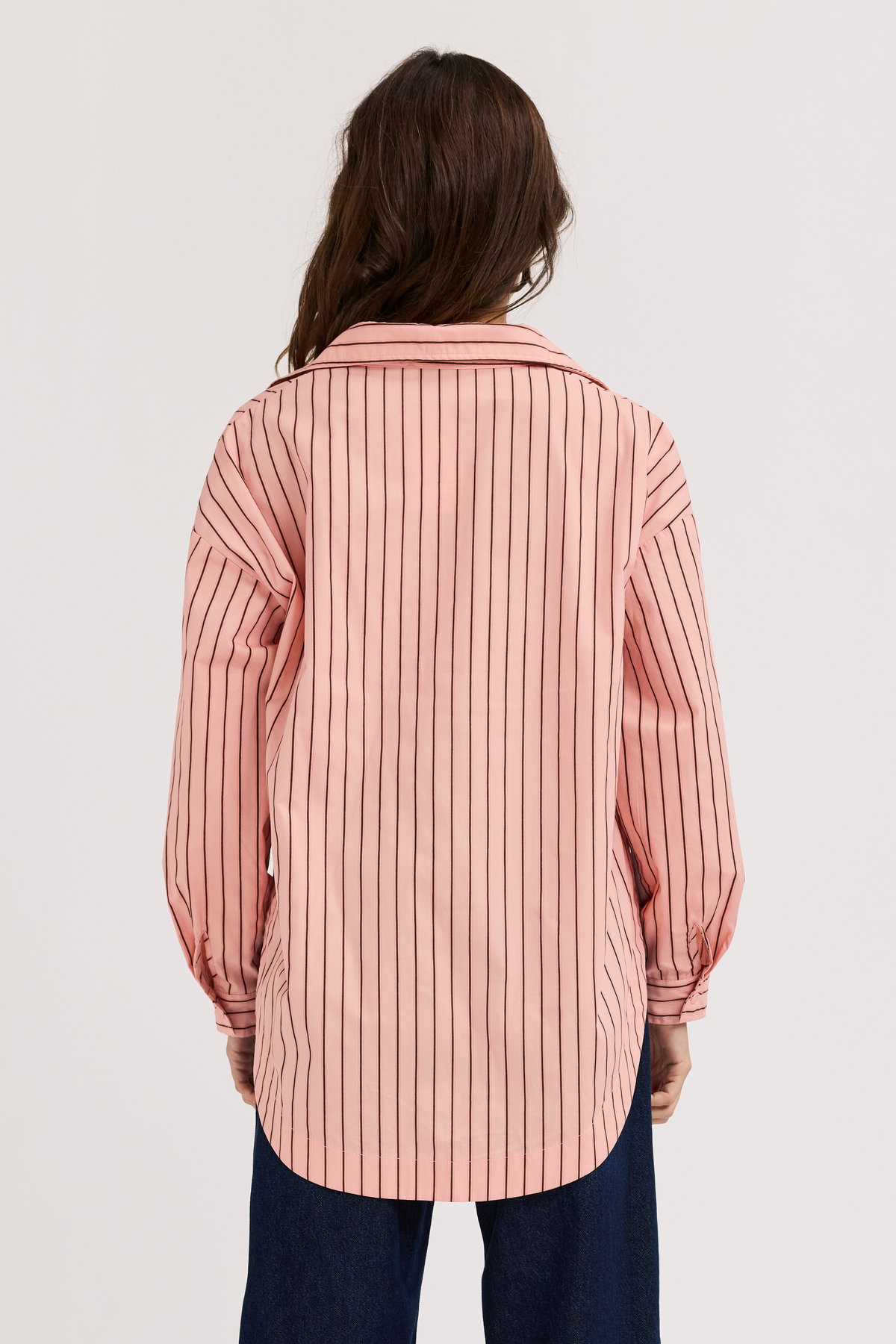 A woman viewed from behind wearing a pink button-up shirt with thin vertical dark stripes and dark blue jeans, set against a plain light gray studio background.